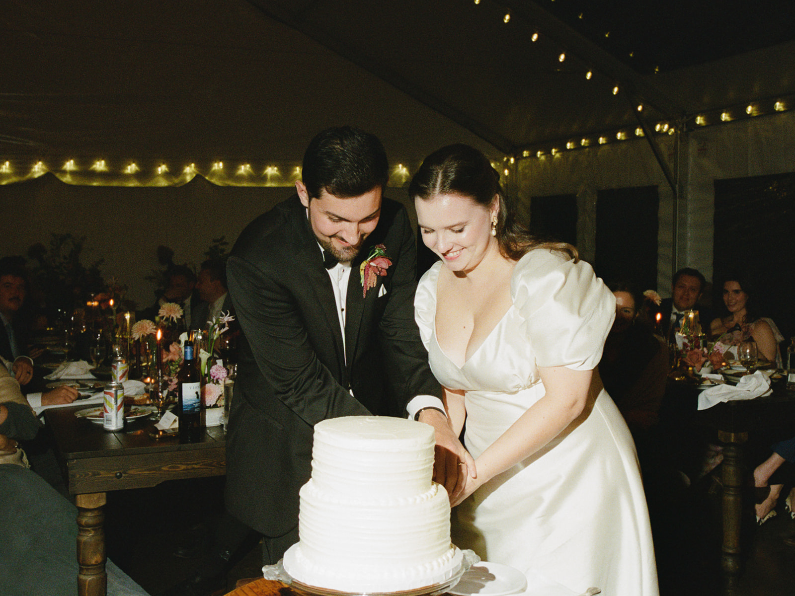 Couple cutting wedding cake under string lights at tented reception